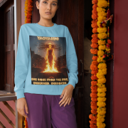 A young woman, possibly of South Asian descent, with long dark hair, wearing a baby blue PodLyft Yagyaseni Fireborn sweatshirt. The sweatshirt features a stylized graphic of a woman's silhouette against a fiery, abstract background, with the text "YAGYASENI" written below. She is looking directly at the camera with a confident expression, her arms crossed, standing against a plain light grey background.