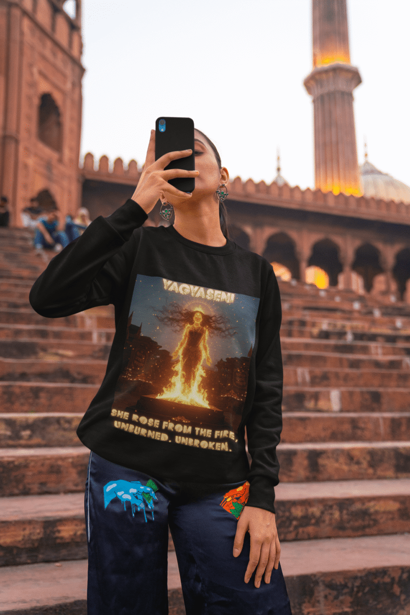 A young woman, possibly of South Asian descent, with long dark hair, wearing a black PodLyft Yagyaseni Fireborn sweatshirt. The sweatshirt features a stylized graphic of a woman's silhouette against a fiery, abstract background, with the text "YAGYASENI" written below. She is looking directly at the camera with a confident expression, her arms crossed, standing against a plain light grey background.