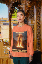 A young woman, possibly of South Asian descent, with long dark hair, wearing a coral PodLyft Yagyaseni Fireborn sweatshirt. The sweatshirt features a stylized graphic of a woman's silhouette against a fiery, abstract background, with the text "YAGYASENI" written below. She is looking directly at the camera with a confident expression, her arms crossed, standing against a plain light grey background.