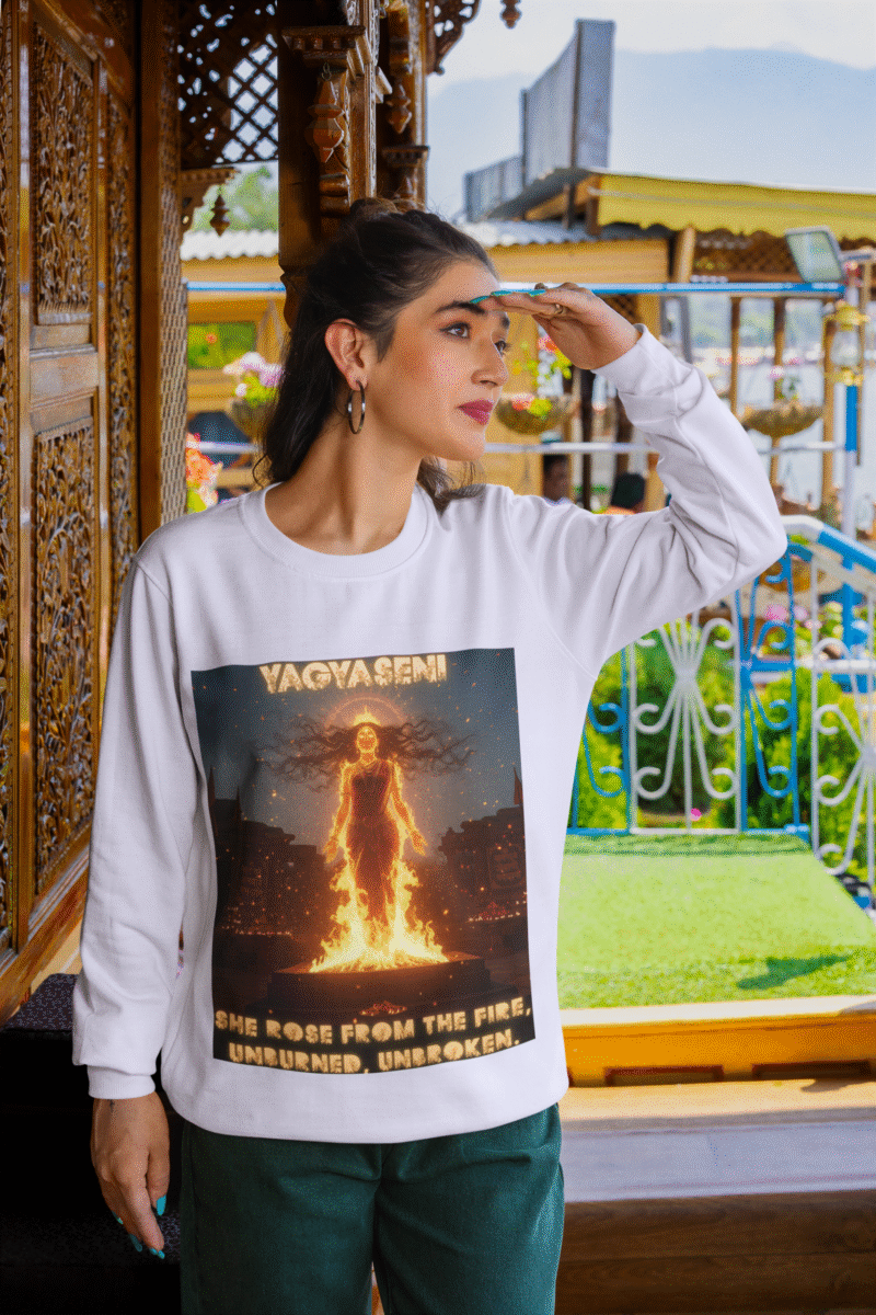 A young woman, possibly of South Asian descent, with long dark hair, wearing a lavender PodLyft Yagyaseni Fireborn sweatshirt. The sweatshirt features a stylized graphic of a woman's silhouette against a fiery, abstract background, with the text "YAGYASENI" written below. She is looking directly at the camera with a confident expression, her arms crossed, standing against a plain light grey background.