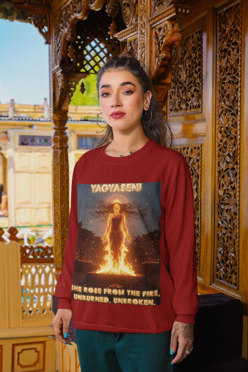 A young woman, possibly of South Asian descent, with long dark hair, wearing a maroon PodLyft Yagyaseni Fireborn sweatshirt. The sweatshirt features a stylized graphic of a woman's silhouette against a fiery, abstract background, with the text "YAGYASENI" written below. She is looking directly at the camera with a confident expression, her arms crossed, standing against a plain light grey background.