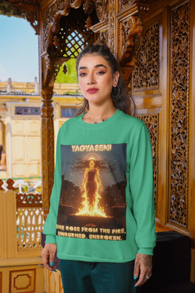 A young woman, possibly of South Asian descent, with long dark hair, wearing a mint green PodLyft Yagyaseni Fireborn sweatshirt. The sweatshirt features a stylized graphic of a woman's silhouette against a fiery, abstract background, with the text "YAGYASENI" written below. She is looking directly at the camera with a confident expression, her arms crossed, standing against a plain light grey background.