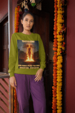 A young woman, possibly of South Asian descent, with long dark hair, wearing an olive green PodLyft Yagyaseni Fireborn sweatshirt. The sweatshirt features a stylized graphic of a woman's silhouette against a fiery, abstract background, with the text "YAGYASENI" written below. She is looking directly at the camera with a confident expression, her arms crossed, standing against a plain light grey background.