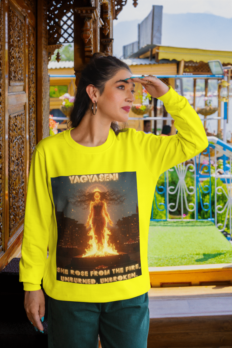 A young woman, possibly of South Asian descent, with long dark hair, wearing a yellow PodLyft Yagyaseni Fireborn sweatshirt. The sweatshirt features a stylized graphic of a woman's silhouette against a fiery, abstract background, with the text "YAGYASENI" written below. She is looking directly at the camera with a confident expression, her arms crossed, standing against a plain light grey background.