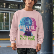 A young man, possibly of Caucasian descent, with short dark hair and a beard, wearing a baby pink PodLyft Mental Wellness Sweatshirt for men. The sweatshirt features a circular graphic with the letters "BKTYM" prominently in the center, surrounded by the phrase "BE KIND TO YOUR MIND." He is looking directly at the camera with a confident expression, standing against a plain light grey background.
