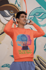 A young man, possibly of Caucasian descent, with short dark hair and a beard, wearing a coral PodLyft Mental Wellness Sweatshirt for men. The sweatshirt features a circular graphic with the letters "BKTYM" prominently in the center, surrounded by the phrase "BE KIND TO YOUR MIND." He is looking directly at the camera with a confident expression, standing against a plain light grey background.