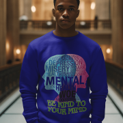 A young man, possibly of Caucasian descent, with short dark hair and a beard, wearing a navy blue PodLyft Mental Wellness Sweatshirt for men. The sweatshirt features a circular graphic with the letters "BKTYM" prominently in the center, surrounded by the phrase "BE KIND TO YOUR MIND." He is looking directly at the camera with a confident expression, standing against a plain light grey background.