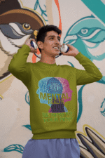 A young man, possibly of Caucasian descent, with short dark hair and a beard, wearing an olive green PodLyft Mental Wellness Sweatshirt for men. The sweatshirt features a circular graphic with the letters "BKTYM" prominently in the center, surrounded by the phrase "BE KIND TO YOUR MIND." He is looking directly at the camera with a confident expression, standing against a plain light grey background.