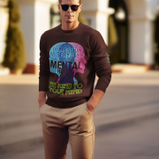 A young man, possibly of Caucasian descent, with short dark hair and a beard, wearing a reddish-brown PodLyft Mental Wellness Sweatshirt for men. The sweatshirt features a circular graphic with the letters "BKTYM" prominently in the center, surrounded by the phrase "BE KIND TO YOUR MIND." He is looking directly at the camera with a confident expression, standing against a plain light grey background.