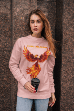A young woman, possibly of Caucasian descent, with light brown hair pulled back, wearing a baby pink PodLyft Phoenix Reign sweatshirt. The sweatshirt features a graphic of a stylized, fiery phoenix with the text "I BURN I RISE I REIGN" below it. She is looking directly at the camera with a confident expression, standing against a plain light grey background.