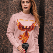 A young woman, possibly of Caucasian descent, with light brown hair pulled back, wearing a baby pink PodLyft Phoenix Reign sweatshirt. The sweatshirt features a graphic of a stylized, fiery phoenix with the text "I BURN I RISE I REIGN" below it. She is looking directly at the camera with a confident expression, standing against a plain light grey background.