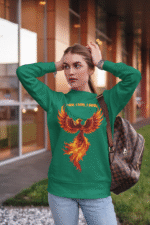 A young woman, possibly of Caucasian descent, with light brown hair pulled back, wearing a bottle green PodLyft Phoenix Reign sweatshirt. The sweatshirt features a graphic of a stylized, fiery phoenix with the text "I BURN I RISE I REIGN" below it. She is looking directly at the camera with a confident expression, standing against a plain light grey background.