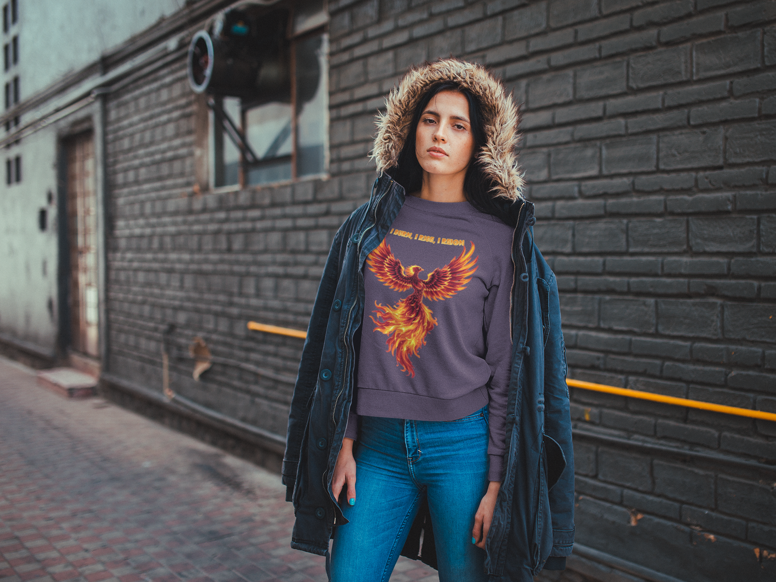 A young woman, possibly of Caucasian descent, with light brown hair pulled back, wearing a brown PodLyft Phoenix Reign sweatshirt. The sweatshirt features a graphic of a stylized, fiery phoenix with the text "I BURN I RISE I REIGN" below it. She is standing outdoors with a confident expression, looking into the distance, against a blurred background of a building facade.