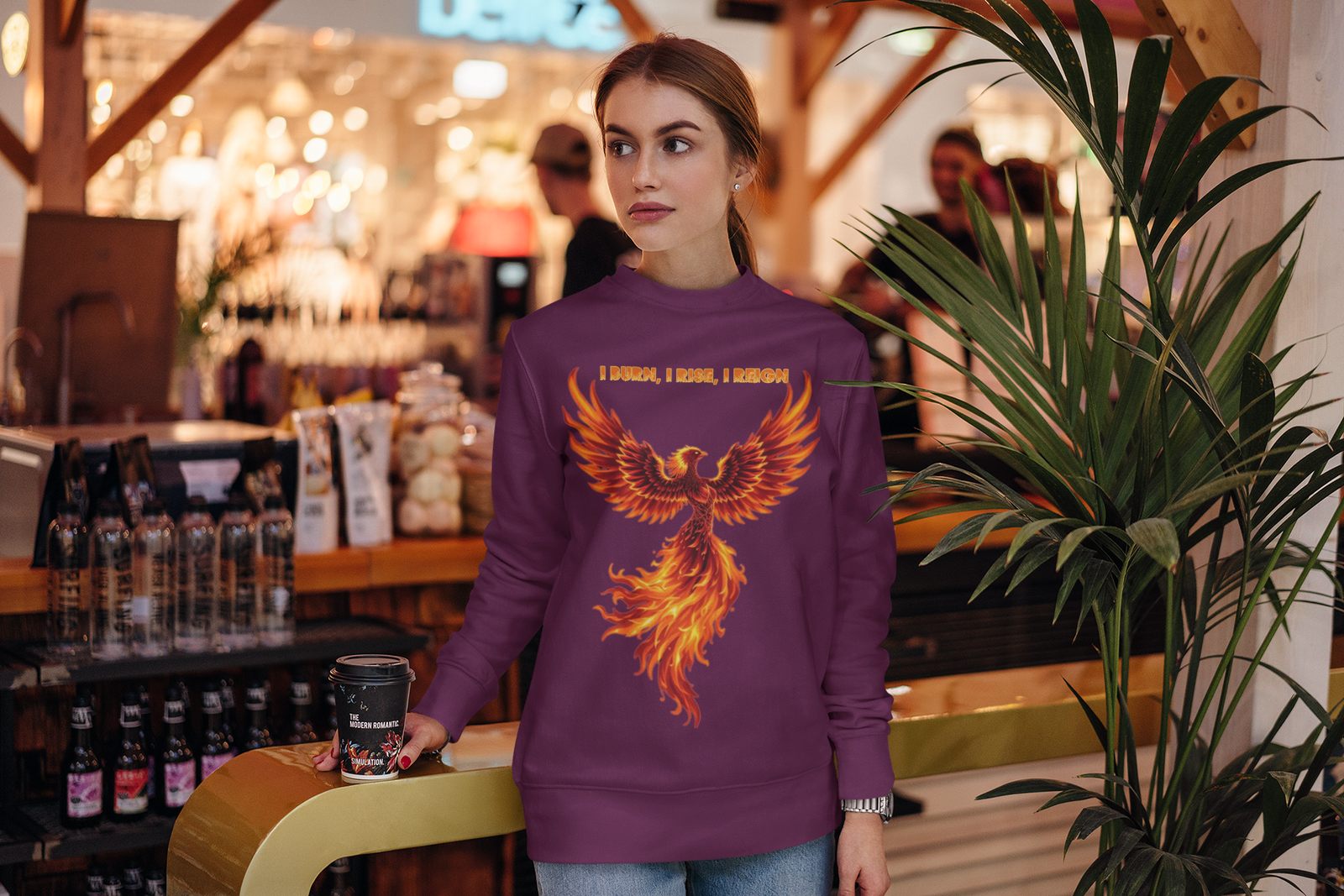 A young woman, possibly of Caucasian descent, with light brown hair pulled back, wearing a purple PodLyft Phoenix Reign sweatshirt. The sweatshirt features a graphic of a stylized, fiery phoenix with the text "I BURN I RISE I REIGN" below it. She is smiling and looking to her left, standing in what appears to be a dimly lit, possibly urban or industrial, setting.