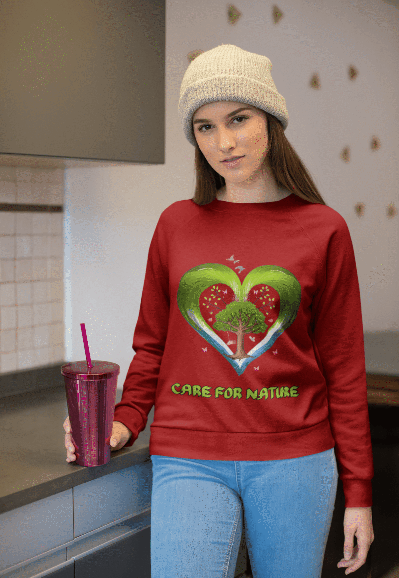 A young woman, possibly of Caucasian descent, with light brown hair, wearing a maroon PodLyft Women's Eco Love Sweatshirt. The sweatshirt features a circular graphic with the phrase "CARE FOR NATURE" prominently displayed. She is looking directly at the camera with a confident yet gentle expression, standing against a plain light grey background.