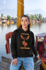A young woman, possibly of Caucasian descent, with light brown hair pulled back, wearing a black PodLyft Women's Revolution Hoodie. The hoodie features a graphic of a stylized, strong female figure, with the text "I AM THE FIRE I AM THE REVOLUTION" below it. She is looking directly at the camera with a confident expression, standing against a plain light grey background.