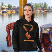 A young woman, possibly of Caucasian descent, with light brown hair pulled back, wearing a black PodLyft Women's Revolution Hoodie. The hoodie features a graphic of a stylized, strong female figure, with the text "I AM THE FIRE I AM THE REVOLUTION" below it. She is looking directly at the camera with a confident expression, standing against a plain light grey background.