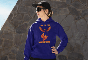 A young woman, possibly of Caucasian descent, with light brown hair, wearing a navy blue PodLyft Women's Revolution Hoodie. The hoodie features a graphic of a stylized, strong female figure, with the text "I AM THE FIRE I AM THE REVOLUTION" below it. She is standing outdoors with a confident expression, looking to her right, against a blurred urban background with green foliage.
