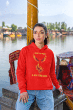 A young woman, possibly of Caucasian descent, with light brown hair pulled back, wearing a red PodLyft Women's Revolution Hoodie. The hoodie features a graphic of a stylized, strong female figure, with the text "I AM THE FIRE I AM THE REVOLUTION" below it. She is looking directly at the camera with a confident expression, standing against a plain light grey background.