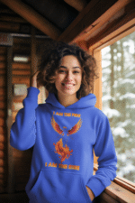 A young woman, possibly of Caucasian descent, with light brown hair pulled back, wearing a royal blue PodLyft Women's Revolution Hoodie. The hoodie features a graphic of a stylized, strong female figure, with the text "I AM THE FIRE I AM THE REVOLUTION" below it. She is looking directly at the camera with a confident expression, standing against a plain light grey background.