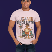A young man, possibly of Caucasian descent, with short dark hair and a beard, wearing a light grayish orange PodLyft Men's Jugaadu t-shirt. The t-shirt features a graphic with the word "JUGAADU SINCE BIRTH." He is looking directly at the camera with a confident expression, standing against a plain light grey background.