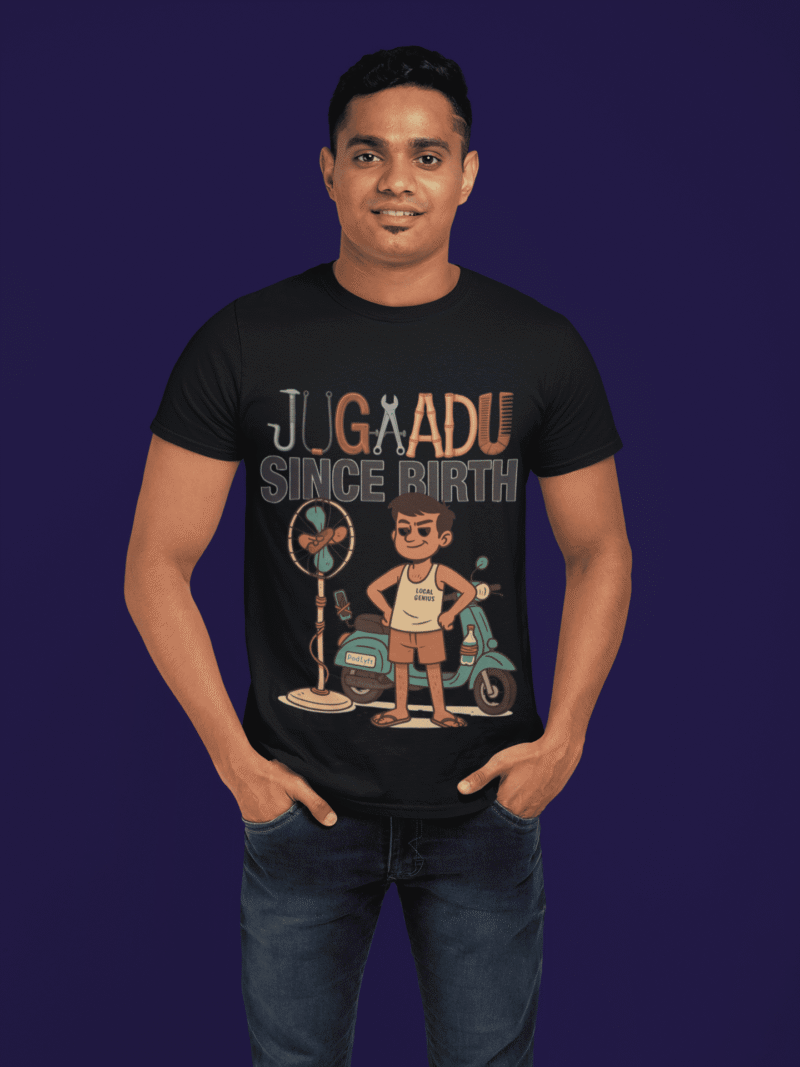 A young man, possibly of Caucasian descent, with short dark hair and a beard, wearing a black PodLyft Men's Jugaadu t-shirt. The t-shirt features a graphic with the text "JUGAADU SINCE BIRTH." He is looking directly at the camera with a confident expression, standing against a plain light grey background.