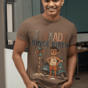 A young man, possibly of Caucasian descent, with short dark hair and a beard, wearing a coffee brown PodLyft Men's Jugaadu t-shirt. The t-shirt features a graphic with the text "JUGAADU SINCE BIRTH." He is looking directly at the camera with a confident expression, standing against a plain light grey background.