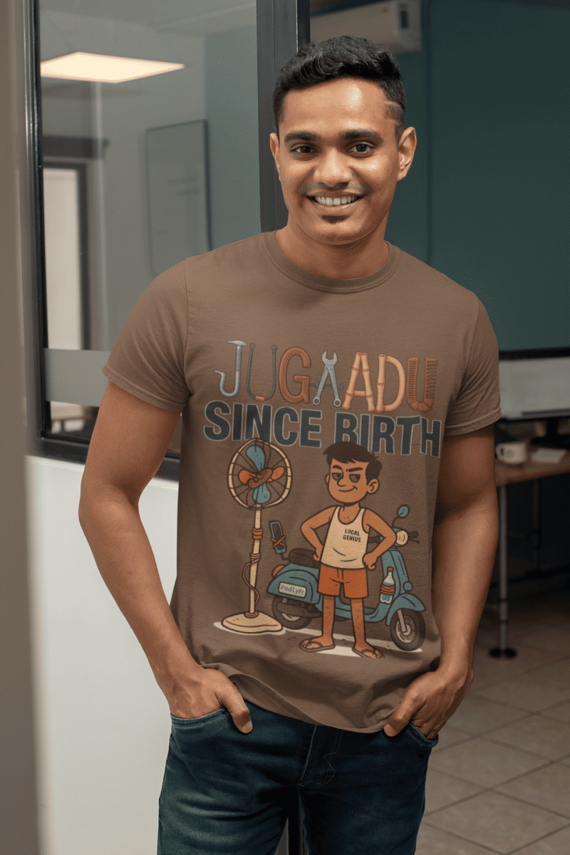 A young man, possibly of Caucasian descent, with short dark hair and a beard, wearing a coffee brown PodLyft Men's Jugaadu t-shirt. The t-shirt features a graphic with the text "JUGAADU SINCE BIRTH." He is looking directly at the camera with a confident expression, standing against a plain light grey background.