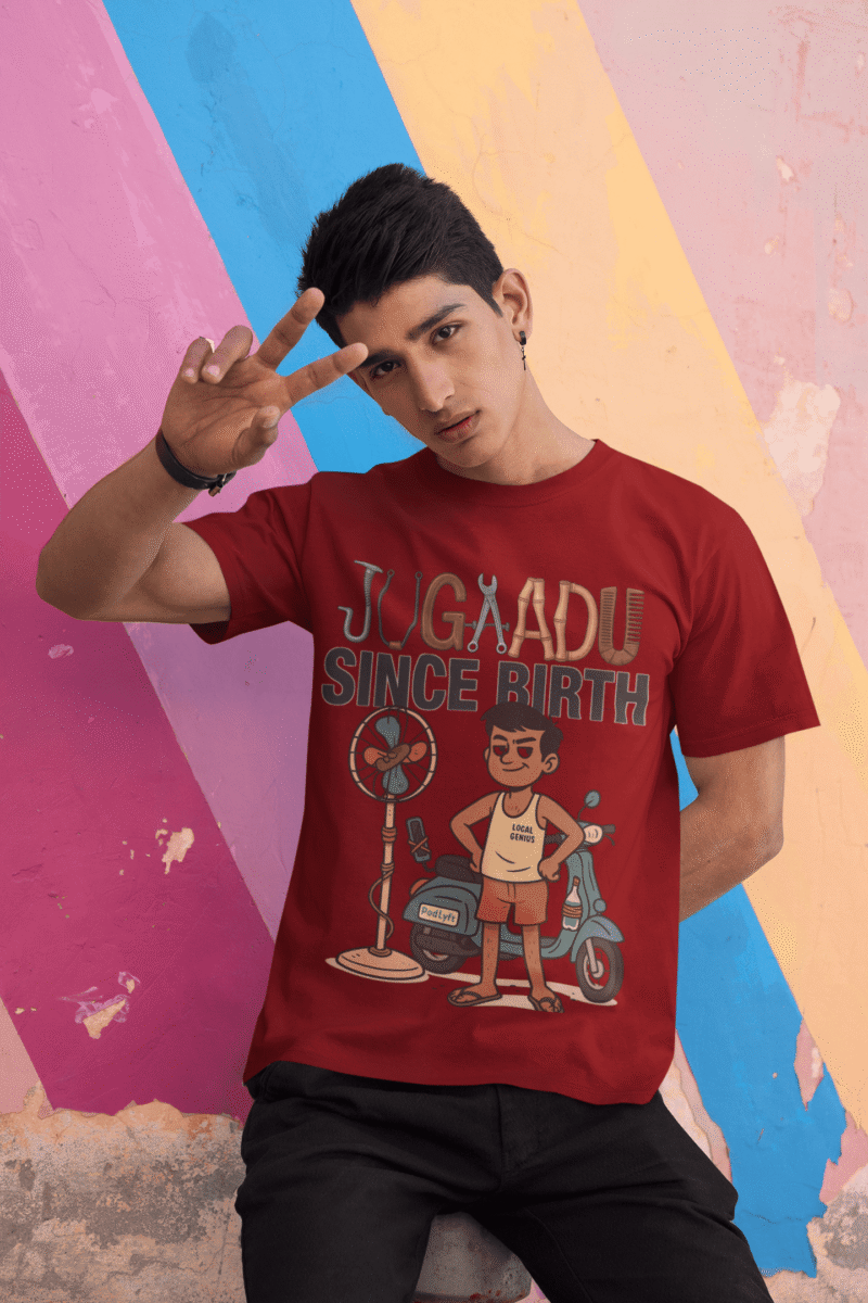 A young man, possibly of Caucasian descent, with short dark hair and a beard, wearing a maroon PodLyft Men's Jugaadu t-shirt. The t-shirt features a graphic with the text "JUGAADU SINCE BIRTH." He is looking directly at the camera with a confident expression, standing against a plain light grey background.