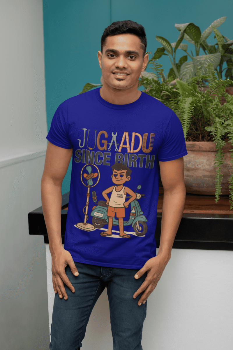 A young man, possibly of Caucasian descent, with short dark hair and a beard, wearing a navy blue PodLyft Men's Jugaadu t-shirt. The t-shirt features a graphic with the text "JUGAADU SINCE BIRTH." He is looking directly at the camera with a confident expression, standing against a plain light grey background.