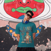 A young man, possibly of Caucasian descent, with short dark hair and a beard, wearing a petrol blue PodLyft Men's Jugaadu t-shirt. The t-shirt features a graphic with the text "JUGAADU SINCE BIRTH." He is looking directly at the camera with a confident expression, standing against a plain light grey background.