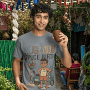 A young man, possibly of Caucasian descent, with short dark hair and a beard, wearing a steel gray PodLyft Men's Jugaadu t-shirt. The t-shirt features a graphic with the text "JUGAADU SINCE BIRTH." He is looking directly at the camera with a confident expression, standing against a plain light grey background.