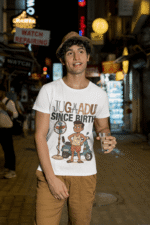 A young man, possibly of Caucasian descent, with short dark hair and a beard, wearing a white PodLyft Men's Jugaadu t-shirt. The t-shirt features a graphic with the text "JUGAADU SINCE BIRTH." He is looking directly at the camera with a confident expression, standing against a plain light grey background.