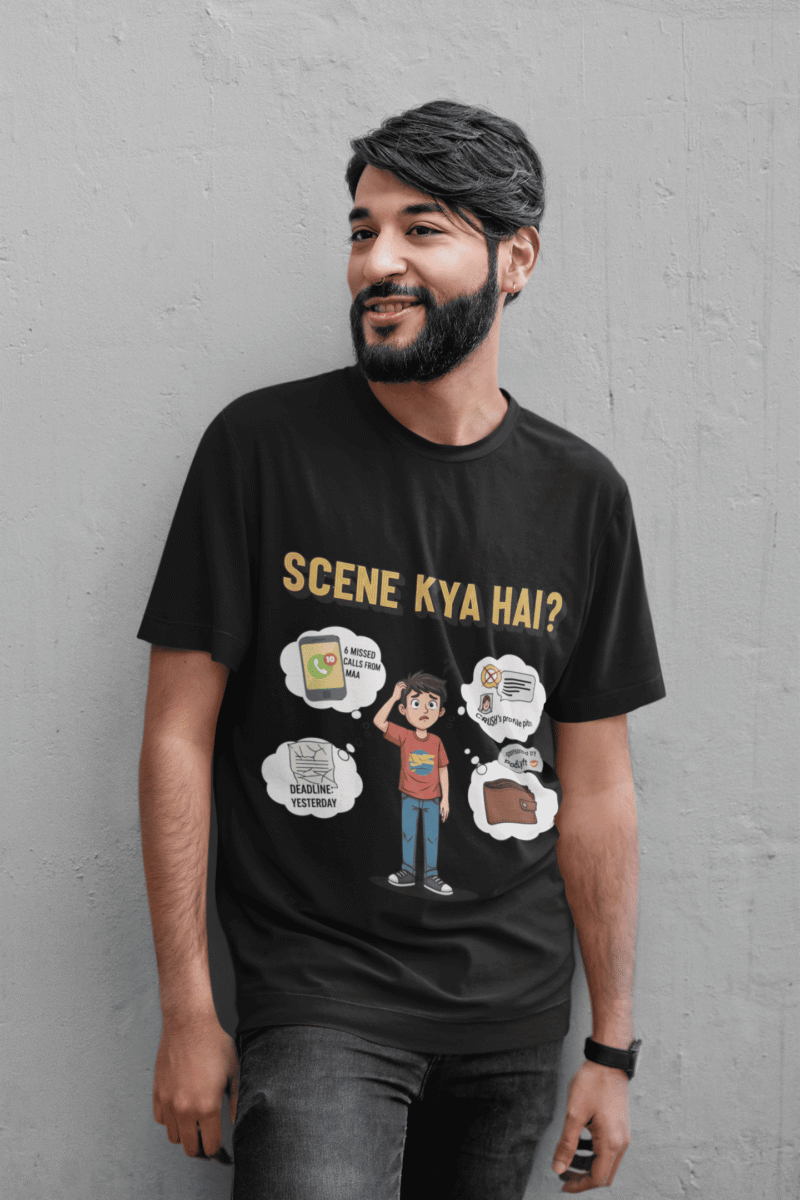 A young man, possibly of Caucasian descent, with short dark hair and a beard, wearing a black t-shirt. The t-shirt features a humorous graphic with the Hindi text "SCENE KYA HAI" and a stylized question mark. He is looking directly at the camera with a confident expression, standing against a plain light grey background.
