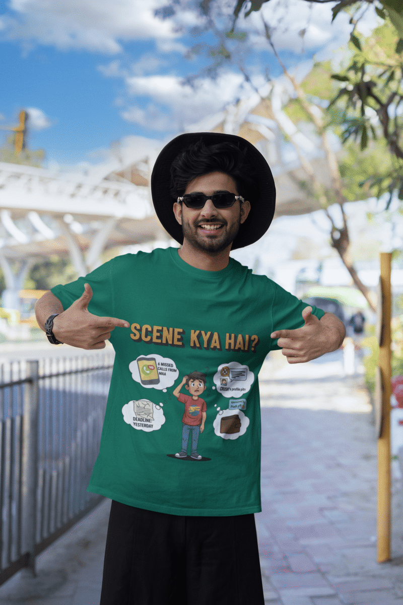 A young man, possibly of Caucasian descent, with short dark hair and a beard, wearing a bottle green t-shirt. The t-shirt features a humorous graphic with the Hindi text "SCENE KYA HAI" and a stylized question mark. He is looking directly at the camera with a confident expression, standing against a plain light grey background.