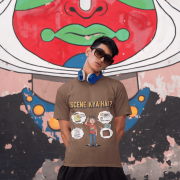 A young man, possibly of Caucasian descent, with short dark hair and a beard, wearing a coffee brown t-shirt. The t-shirt features a humorous graphic with the Hindi text "SCENE KYA HAI" and a stylized question mark. He is looking directly at the camera with a confident expression, standing against a plain light grey background.