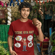 A young man, possibly of Caucasian descent, with short dark hair and a beard, wearing a maroon t-shirt. The t-shirt features a humorous graphic with the Hindi text "SCENE KYA HAI" and a stylized question mark. He is looking directly at the camera with a confident expression, standing against a plain light grey background.
