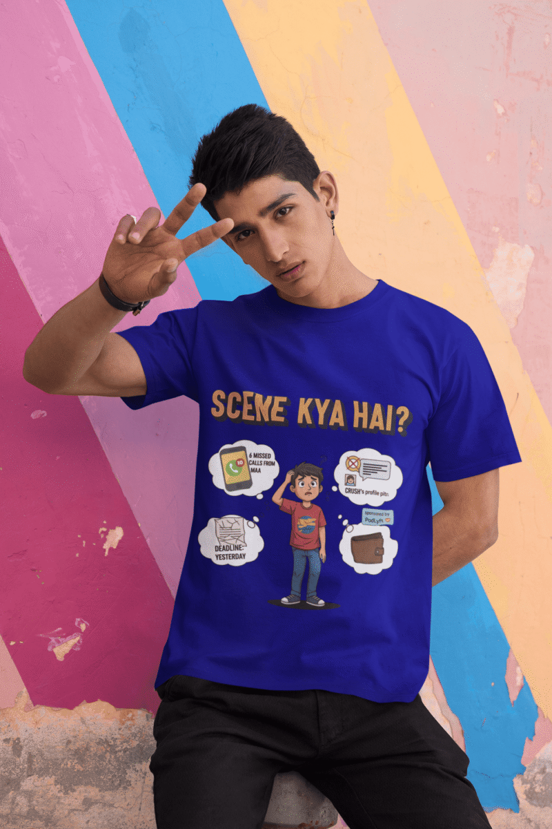 A young man, possibly of Caucasian descent, with short dark hair and a beard, wearing a navy blue t-shirt. The t-shirt features a humorous graphic with the Hindi text "SCENE KYA HAI" and a stylized question mark. He is looking directly at the camera with a confident expression, standing against a plain light grey background.