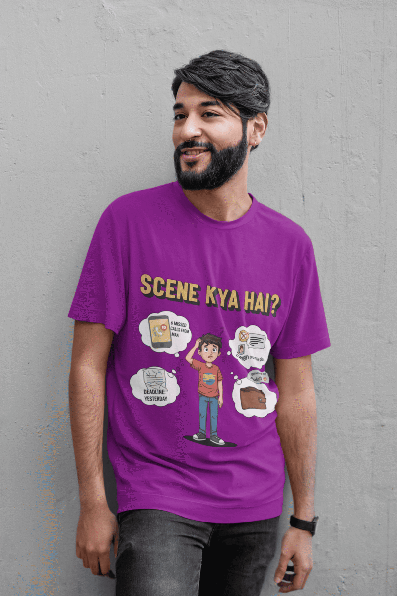 A young man, possibly of Caucasian descent, with short dark hair and a beard, wearing a purple t-shirt. The t-shirt features a humorous graphic with the Hindi text "SCENE KYA HAI" and a stylized question mark. He is looking directly at the camera with a confident expression, standing against a plain light grey background.