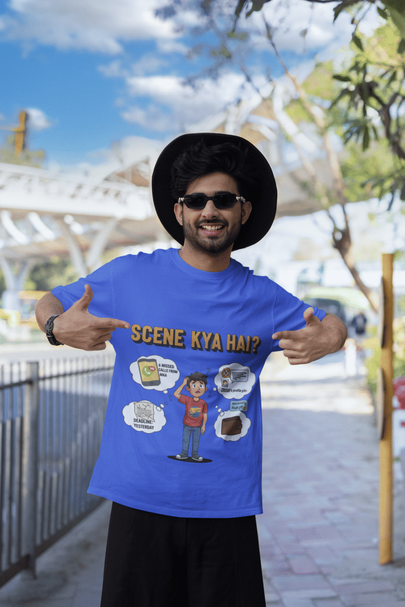 A young man, possibly of Caucasian descent, with short dark hair and a beard, wearing a royal blue t-shirt. The t-shirt features a humorous graphic with the Hindi text "SCENE KYA HAI" and a stylized question mark. He is looking directly at the camera with a confident expression, standing against a plain light grey background.