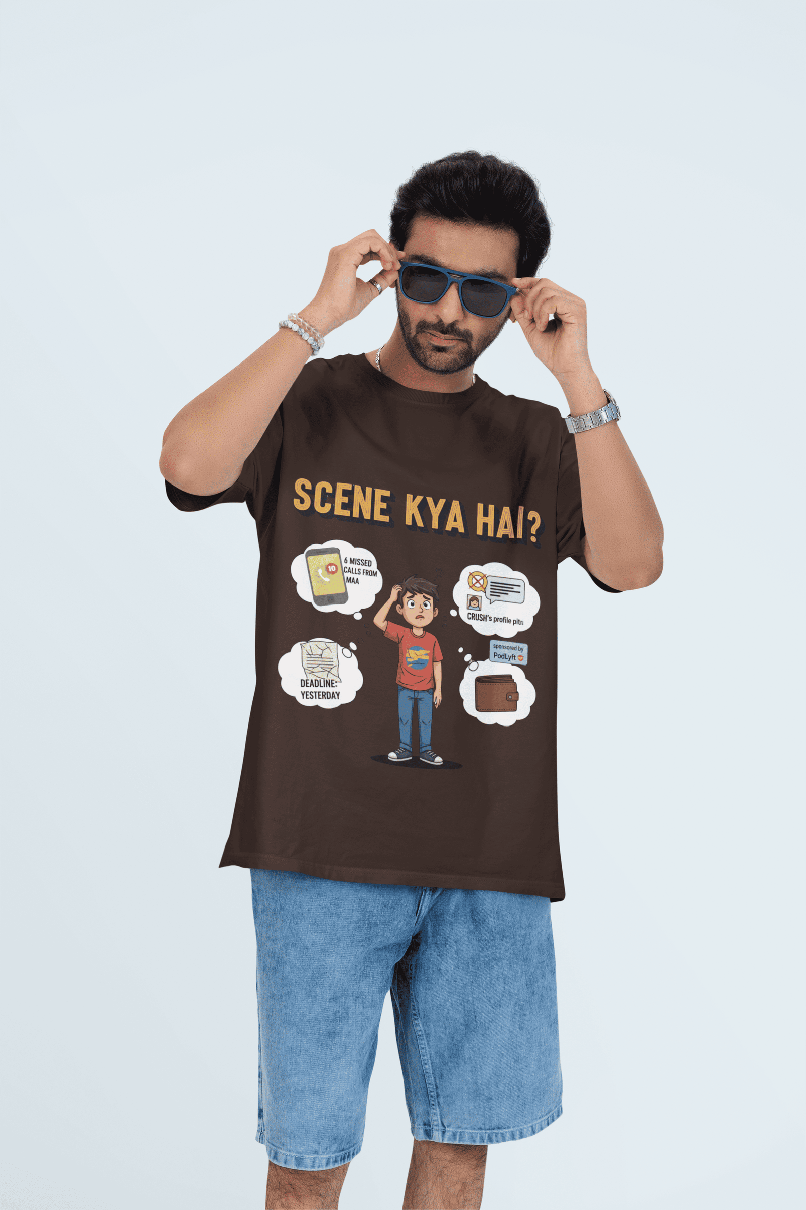 A young man, possibly of Caucasian descent, with short dark hair and a beard, wearing a muted brown Men's Scene Kya Hai T-shirt. The t-shirt features a humorous graphic with the Hindi text "SCENE KYA HAI" and a stylized question mark. He is looking directly at the camera with a confident expression, standing against a plain light grey background.