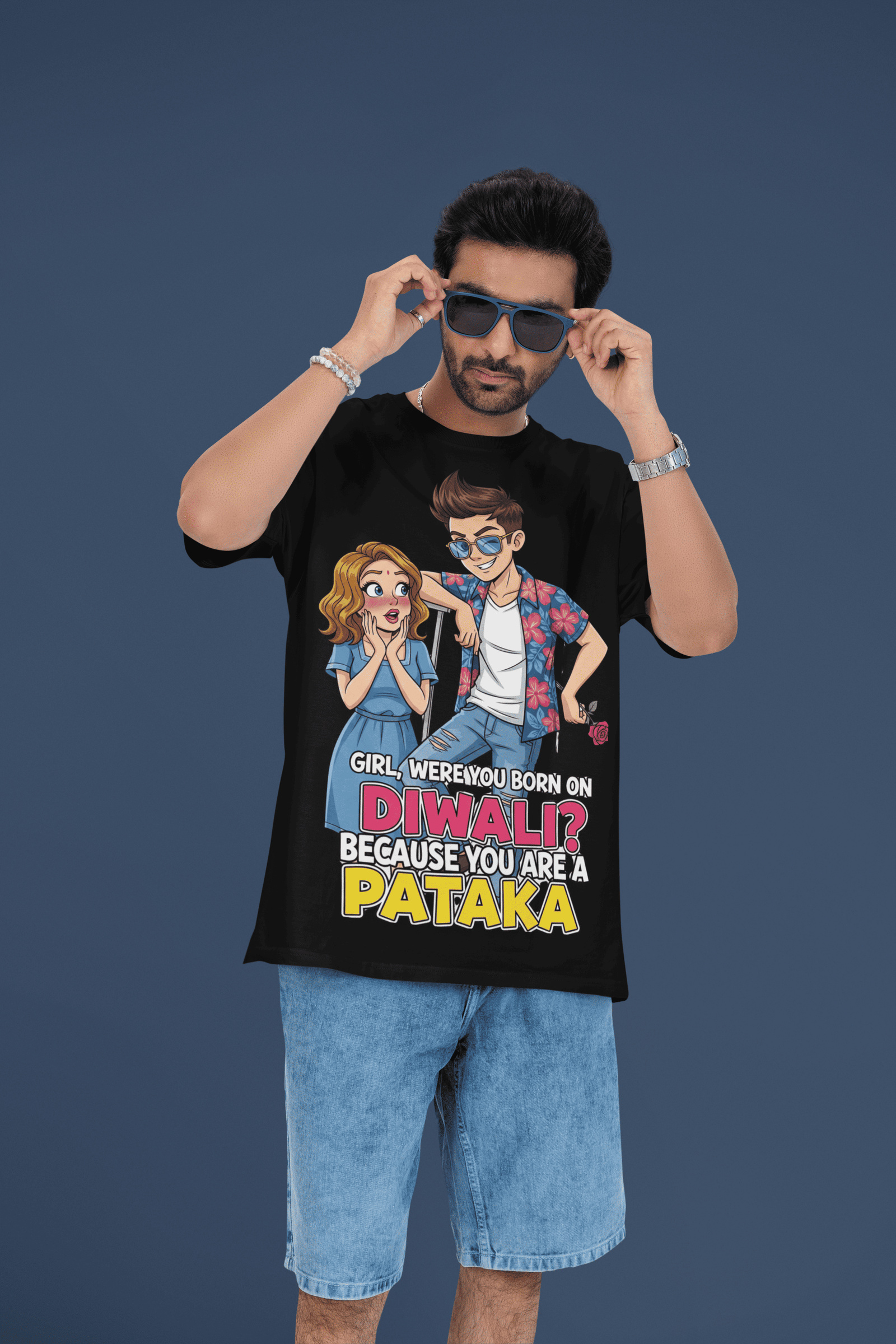 A young man, possibly of Caucasian descent, with short dark hair and a beard, wearing a black PodLyft Pataka t-shirt. The t-shirt features a flirty graphic with the text, "Girl, were you born on Diwali? Because You are PATAKA." He is looking directly at the camera with a confident expression, standing against a plain light grey background.