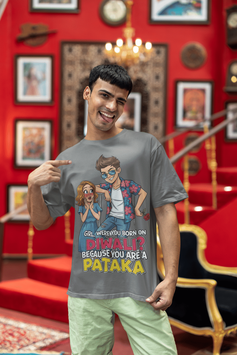 A young man, possibly of Caucasian descent, with short dark hair and a beard, wearing a grey PodLyft Pataka t-shirt. The t-shirt features a flirty graphic with the text, "Girl, were you born on Diwali? Because You are PATAKA." He is looking directly at the camera with a confident expression, standing against a plain light grey background.