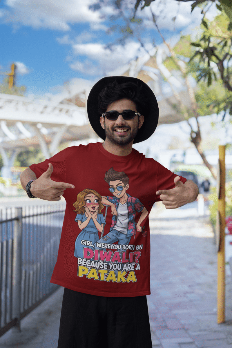 A young man, possibly of Caucasian descent, with short dark hair and a beard, wearing a maroon PodLyft Pataka t-shirt. The t-shirt features a flirty graphic with the text, "Girl, were you born on Diwali? Because You are PATAKA." He is looking directly at the camera with a confident expression, standing against a plain light grey background.