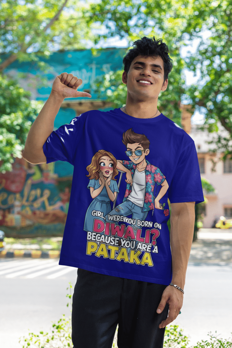 A young man, possibly of Caucasian descent, with short dark hair and a beard, wearing a navy blue PodLyft Pataka t-shirt. The t-shirt features a flirty graphic with the text, "Girl, were you born on Diwali? Because You are PATAKA." He is looking directly at the camera with a confident expression, standing against a plain light grey background.