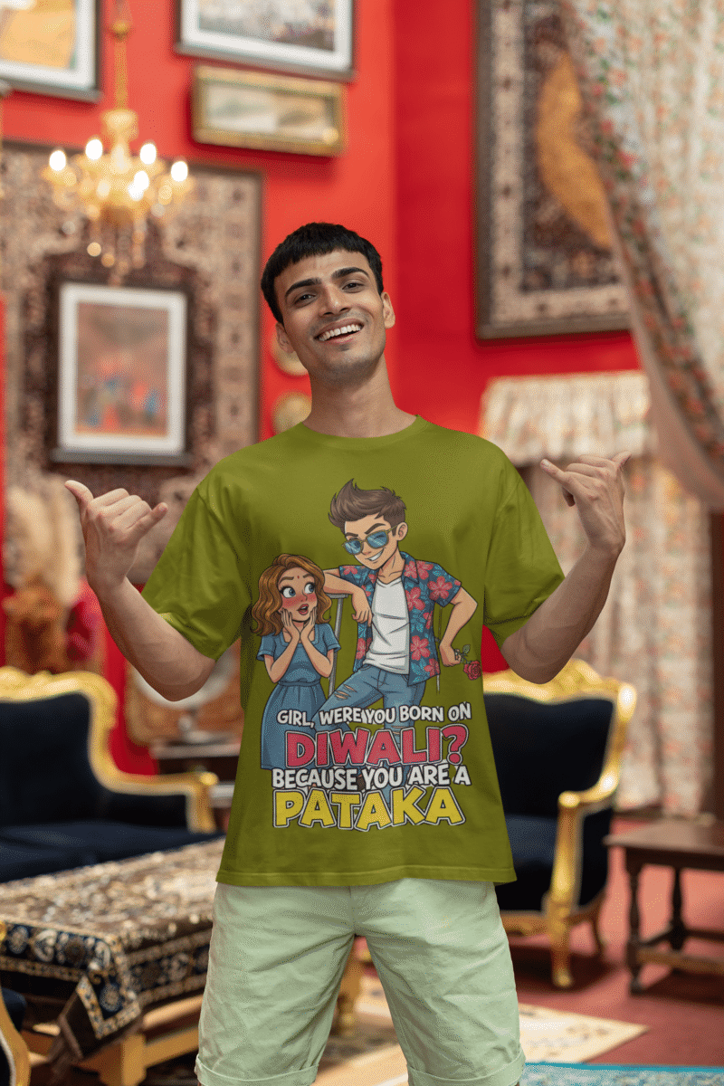 A young man, possibly of Caucasian descent, with short dark hair and a beard, wearing an olive green PodLyft Pataka t-shirt. The t-shirt features a flirty graphic with the text, "Girl, were you born on Diwali? Because You are PATAKA." He is looking directly at the camera with a confident expression, standing against a plain light grey background.
