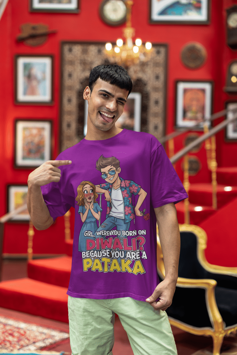A young man, possibly of Caucasian descent, with short dark hair and a beard, wearing a purple PodLyft Pataka t-shirt. The t-shirt features a flirty graphic with the text, "Girl, were you born on Diwali? Because You are PATAKA." He is looking directly at the camera with a confident expression, standing against a plain light grey background.