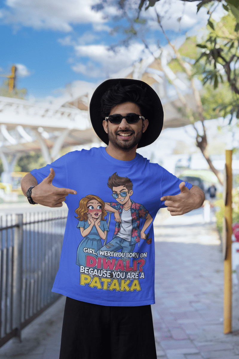 A young man, possibly of Caucasian descent, with short dark hair and a beard, wearing a royal blue PodLyft Pataka t-shirt. The t-shirt features a flirty graphic with the text, "Girl, were you born on Diwali? Because You are PATAKA." He is looking directly at the camera with a confident expression, standing against a plain light grey background.