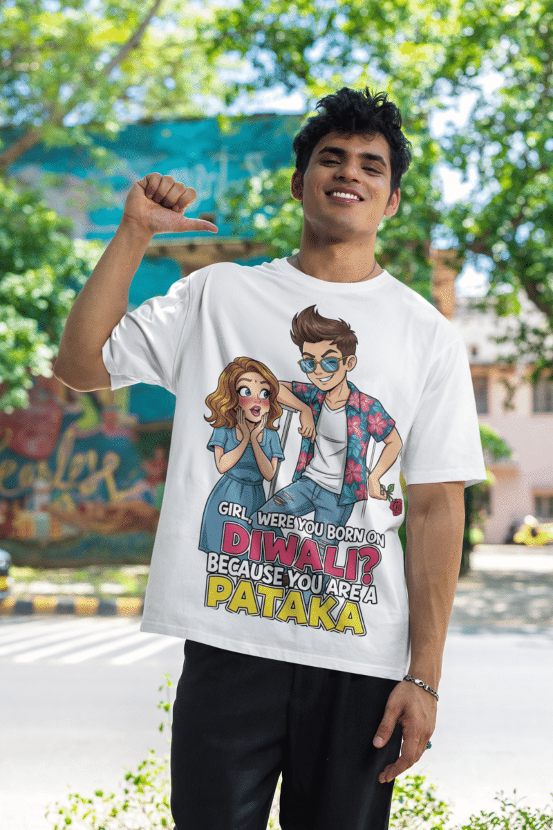 A young man, possibly of Caucasian descent, with short dark hair and a beard, wearing a white PodLyft Pataka t-shirt. The t-shirt features a flirty graphic with the text, "Girl, were you born on Diwali? Because You are PATAKA." He is looking directly at the camera with a confident expression, standing against a plain light grey background.