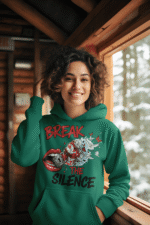 A young woman, possibly of Caucasian descent, with light brown hair pulled back, wearing a bottle green PodLyft Women's Break the Silence Hoodie. The hoodie features a graphic of a padlocked mouth with doves and butterflies bursting out, and the text "BREAK THE SILENCE" and "RAISE YOUR VOICE." She is looking directly at the camera with a confident expression, standing against a plain light grey background.